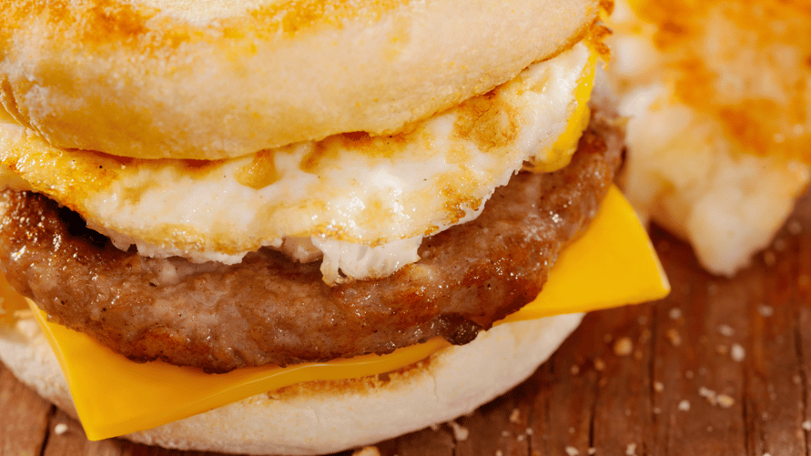 Close up of fried egg, sausage patty and slice of American cheese on toasted English muffin. 