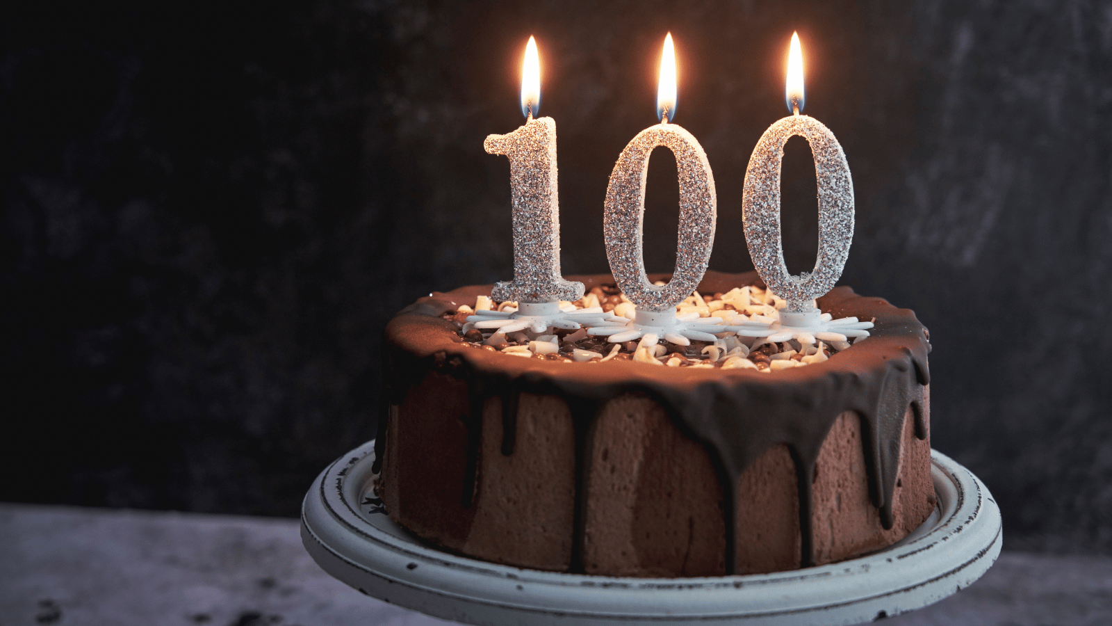 Chocolate frosted cake on cake stand with lit candles that say 100