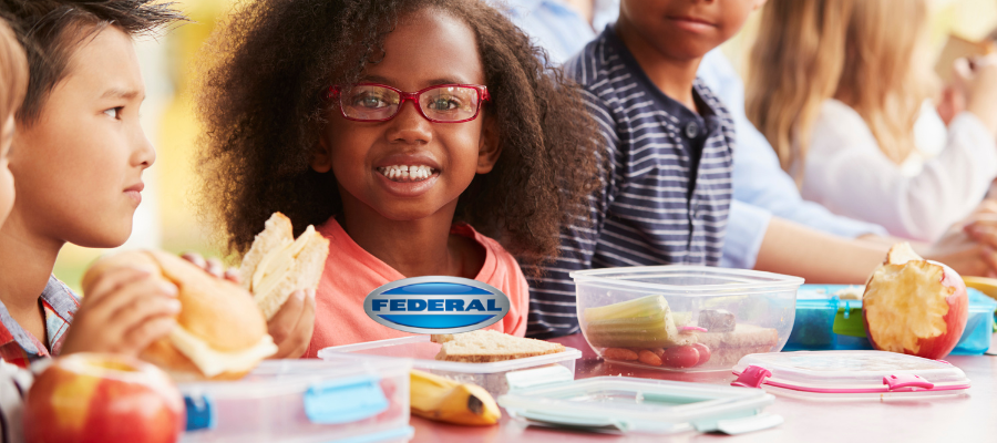 African American girl with curly shoulder length hair and red glasses in school cafeteria looks directly at camera. Other students on either side of her with lunch food in front.