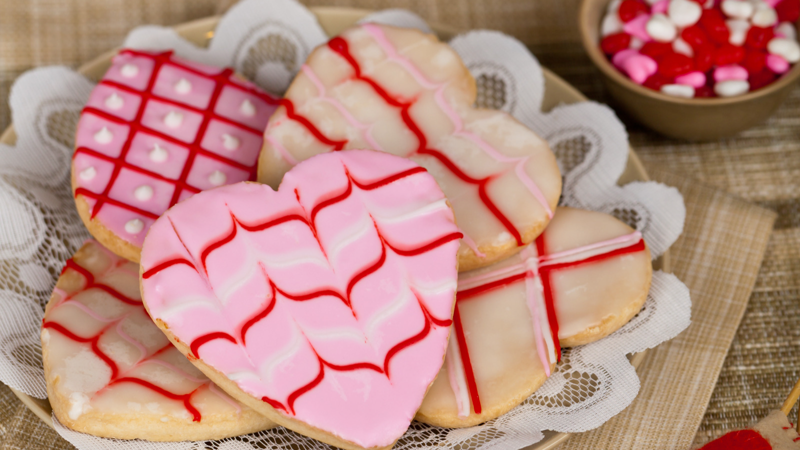 Five heart shaped cookies decorated with pink, white and red frosting on top of doily and plate with heart shaped mini candies in brown bowl behind